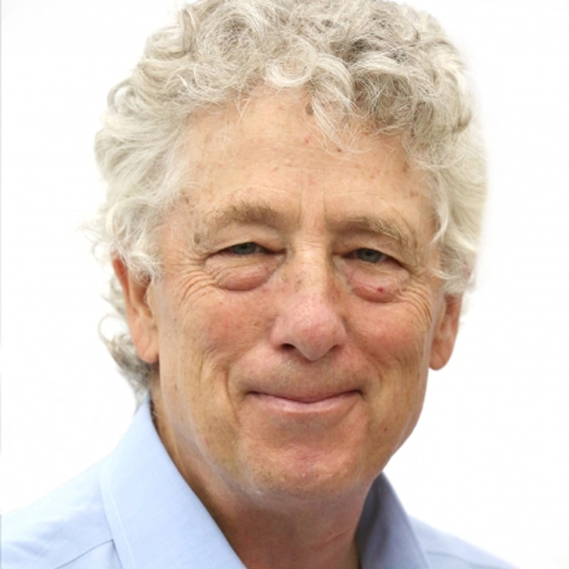 An older man with light curly gray hair and blue eyes smiles gently at the camera. He has fair skin, visible wrinkles, and is wearing a light blue collared shirt. The background is plain white, creating a simple, clean portrait.