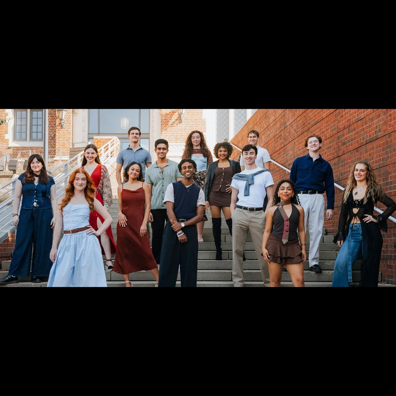 A diverse group of fifteen young adults stands posed on outdoor steps against a brick wall, all smiling and dressed in stylish, semi-formal outfits in earth tones, blues, and whites—proudly representing the CCM Class of 2026 under daylight.