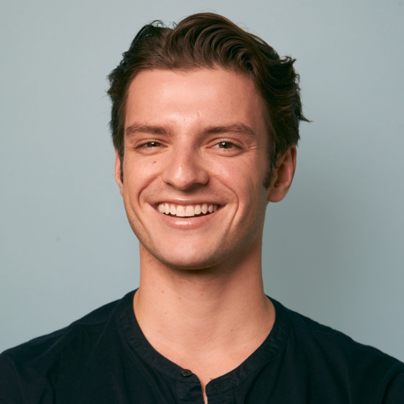 A young man with short brown hair smiles warmly at the camera. He is wearing a dark navy blue shirt with a round neckline. The background is plain light blue, and the lighting is soft, creating a friendly and approachable atmosphere.