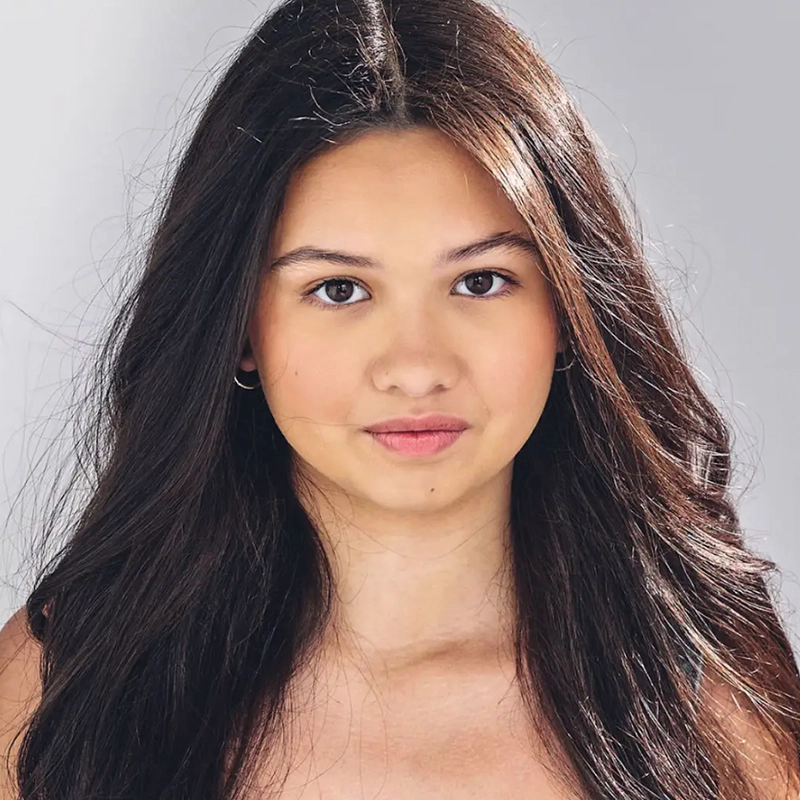 A young woman with long, dark brown hair and light skin looks directly at the camera with a neutral expression. She wears small hoop earrings and has a natural makeup look. The plain, light-colored background highlights Jordan Gulliford's features.