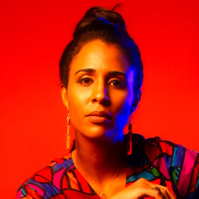 Brianna Rios, with medium skin tone and dark hair in a top bun, poses confidently against a vivid red-orange background. She wears geometric earrings and a colorful top, dramatic blue and yellow lighting highlighting her striking features.