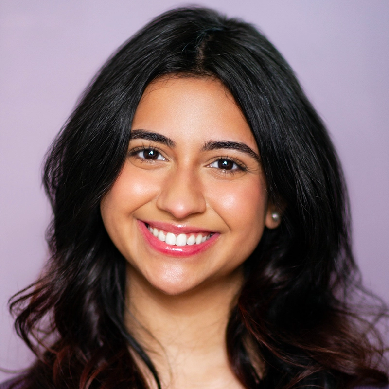 A young woman with long, dark wavy hair smiles warmly at the camera. She has a light to medium skin tone, bright eyes, and glossy pink lips. She is posed in front of a plain, soft purple background.