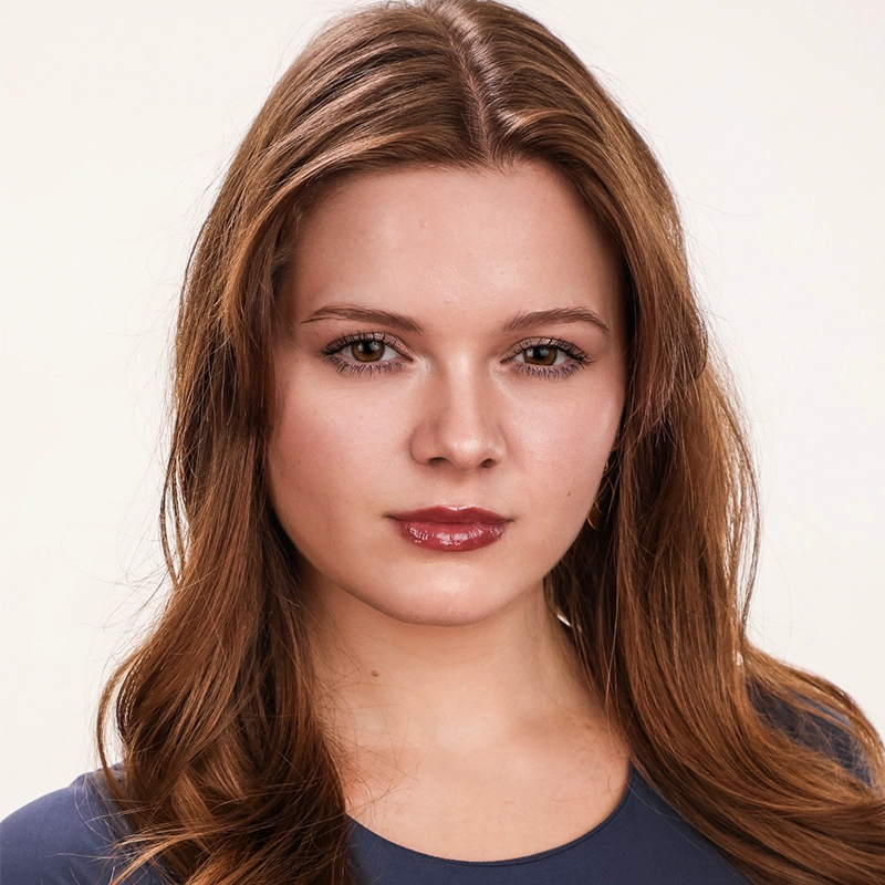A young woman with long, wavy light brown hair and fair skin looks calmly at the camera. She has subtle makeup, glossy lips, and is wearing a blue top. The plain background echoes the minimalist style favored by Jake Hamilton.