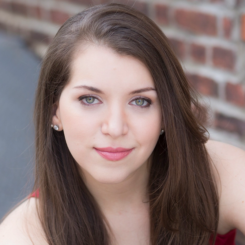 A young woman with long brown hair and light green eyes looks directly at the camera. She has fair skin, is wearing pearl earrings, and a soft pink lipstick. The background is blurred brick and pavement, giving a natural, outdoor setting.