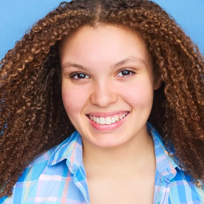 A young person with light brown skin and voluminous curly hair smiles warmly. Jordan Gulliford wears a blue and pink plaid shirt over a white top, set against a solid light blue background that highlights their joyful expression.