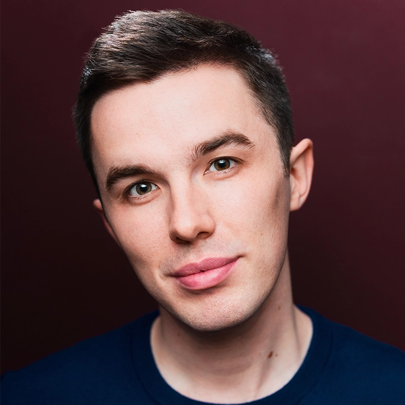 A young man with short brown hair, fair skin, and hazel eyes looks directly at the camera with a neutral expression. He is wearing a dark blue shirt, posed against a solid maroon background in the style of Kristin Weiss.