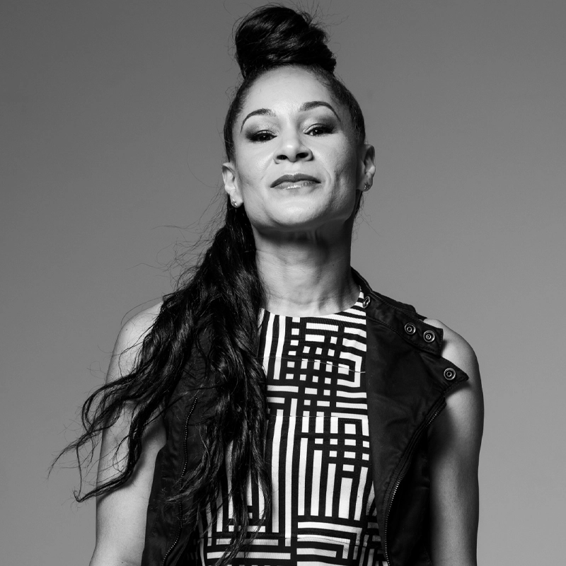 Black and white photo of choreographer Ayodele Casel, a renowned tap dancer, with long, wavy hair in a high topknot. She wears a sleeveless patterned top and dark vest, facing the camera confidently against a plain background.