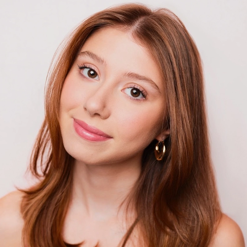 A young woman with straight, shoulder-length light brown hair and fair skin looks at the camera, smiling softly. She wears gold hoop earrings and natural makeup with pink lipstick, in a portrait style reminiscent of Jake Hamilton’s minimal backgrounds.