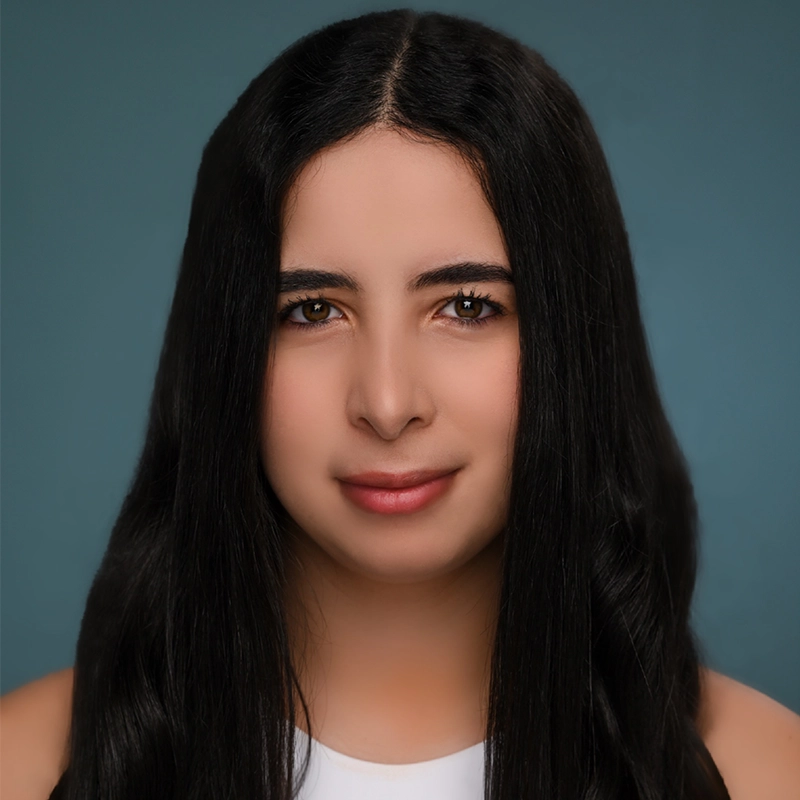 A young woman with long, straight, dark hair poses against a plain teal background. She has light skin, brown eyes, and a subtle smile. She is wearing a sleeveless white top and is looking directly at the camera.