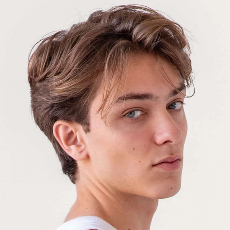 A young person with fair skin, blue eyes, and light brown, wavy hair looks over their shoulder at the camera. With an even complexion and a white shirt, Jordan Gulliford stands against a plain, light-colored background.