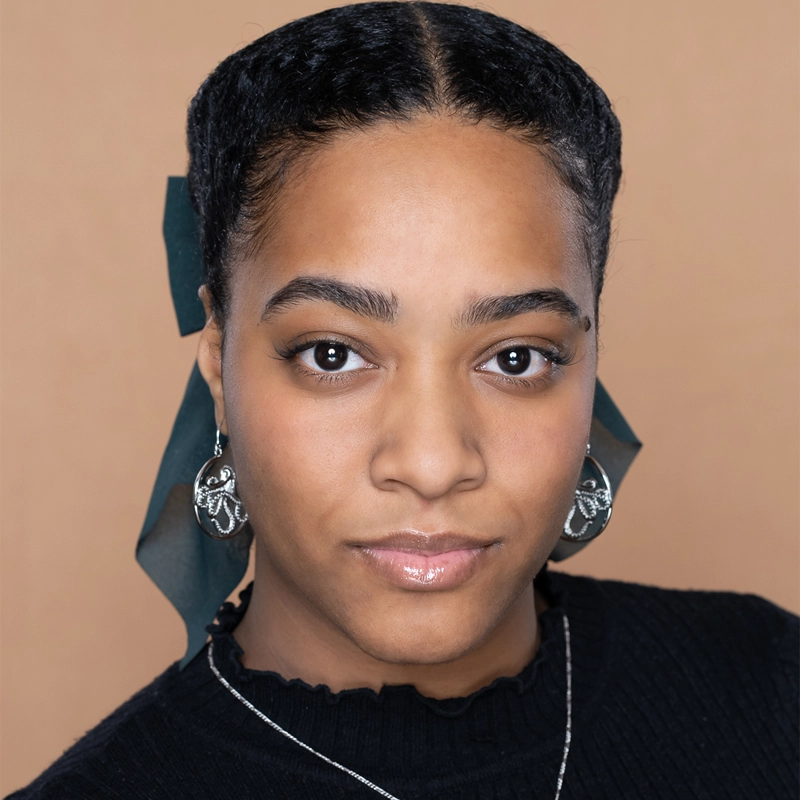 Mariesa Charles, a young woman with medium brown skin and dark braided hair tied with a dark green ribbon, faces the camera. She wears a black top, silver hoop earrings, and a silver necklace against a light brown background.