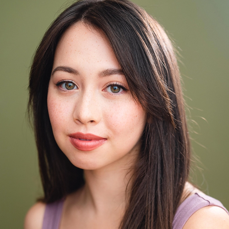 Jenna Leigh Miller, a young woman with fair skin, long straight dark brown hair, and hazel eyes, looks at the camera. She wears natural makeup with pink lipstick and a mauve sleeveless top against a soft, muted green background.