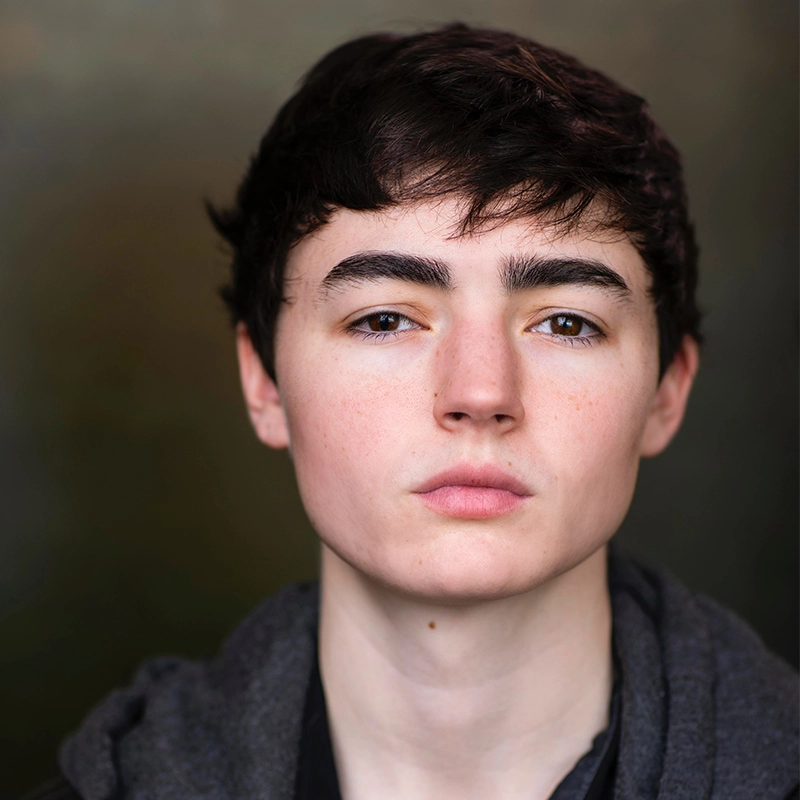 Chris Allyah, a young person with short dark hair and pale skin, looks directly at the camera with a neutral expression. They wear a dark gray hoodie over a black shirt against a softly blurred background of brown and green tones.