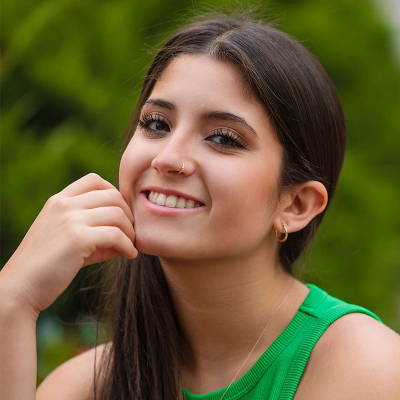Chris Allyah, a young woman with long dark hair, wearing a green sleeveless top, small hoop earrings, and a nose ring, smiles gently at the camera. She rests her hand near her face against a backdrop of blurred greenery.