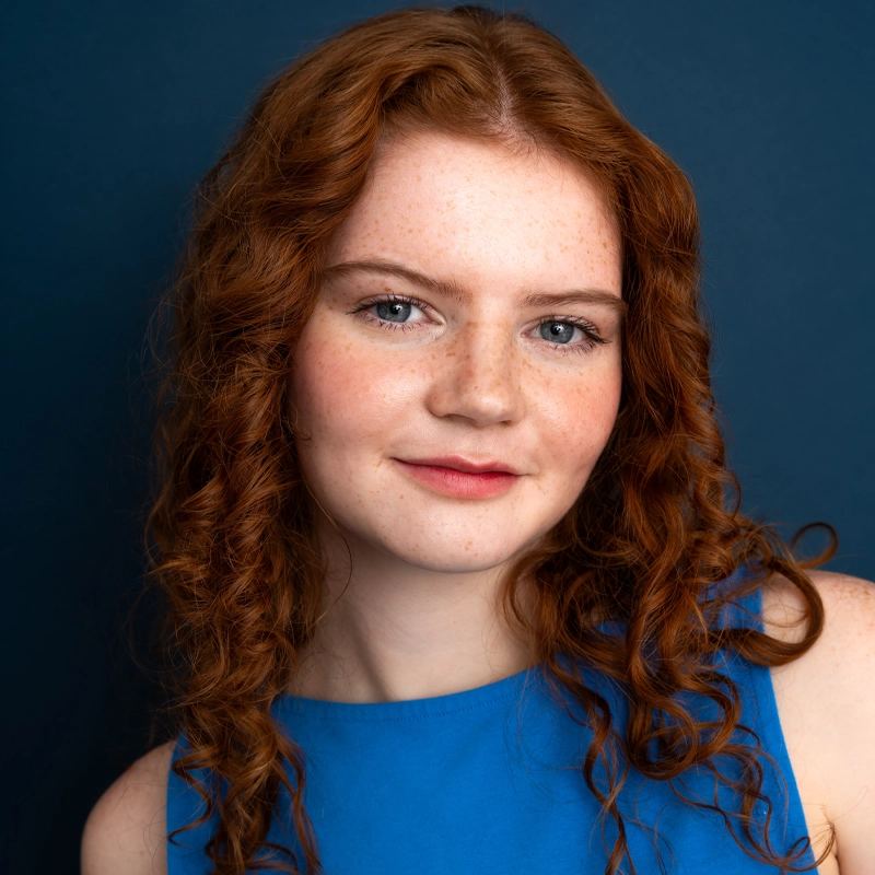A young woman, Mairead Connor, with long, curly red hair and fair, freckled skin smiles softly. She wears a sleeveless royal blue top and poses against a dark blue background, looking directly at the camera with a confident, relaxed expression.