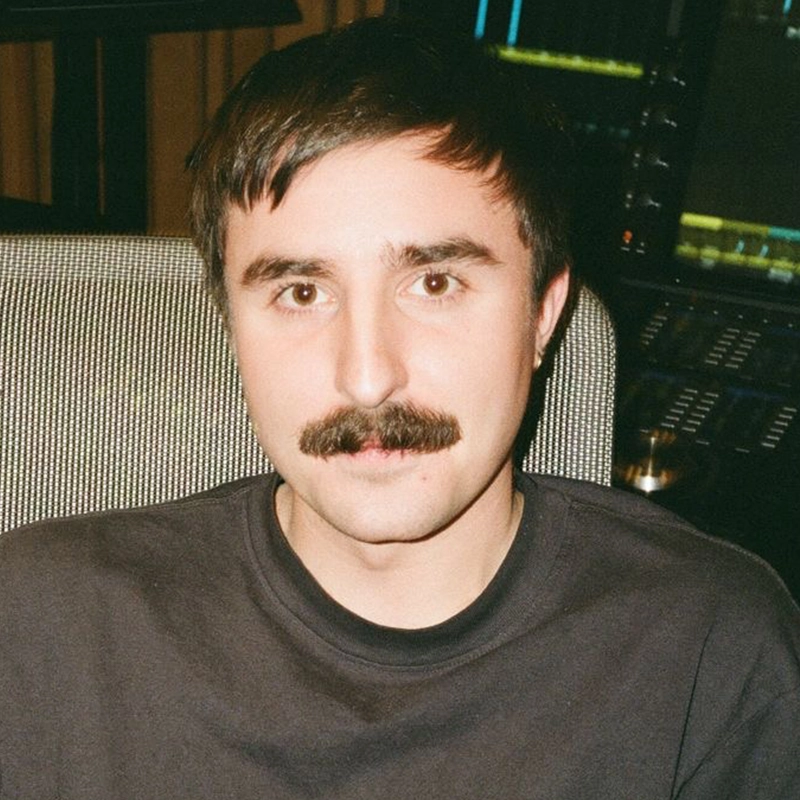 A man with short dark hair and a thick mustache sits in front of audio equipment, wearing a black shirt. He looks at the camera with a neutral expression. The dimly lit background features screens and control panels, hinting at Maya Monteleone's studio.