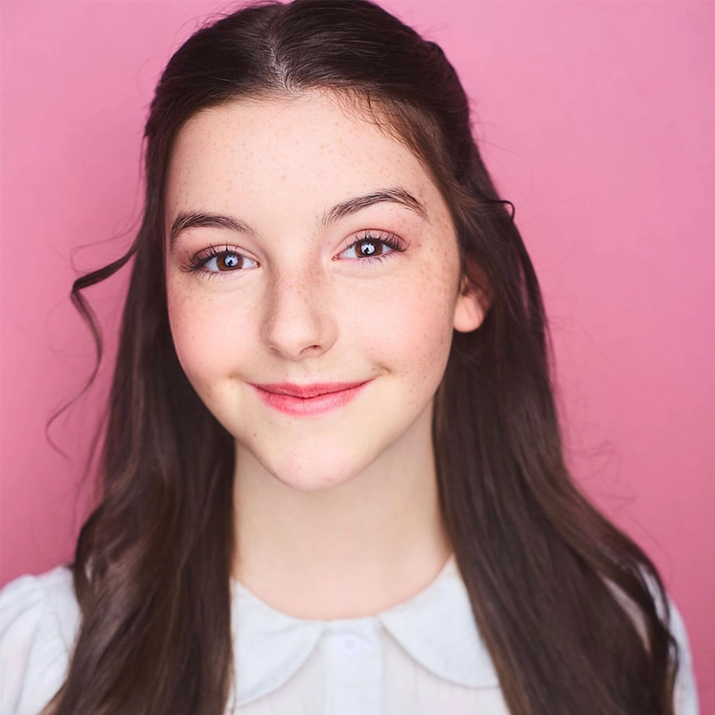 A young woman with fair skin, long dark brown hair, and freckles smiles gently at the camera. She wears a white collared blouse and stands in front of a solid pink background, her style reflecting a classic Matthew Brand aesthetic.