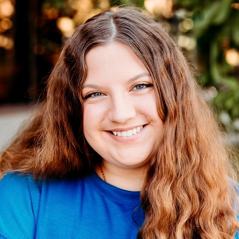A young woman with long, wavy brown hair smiles at the camera. She is wearing a bright blue shirt and is outside, with blurred greenery and sunlight in the background. Her warm, friendly expression captures Gina Marie Incandela’s vibrant spirit.