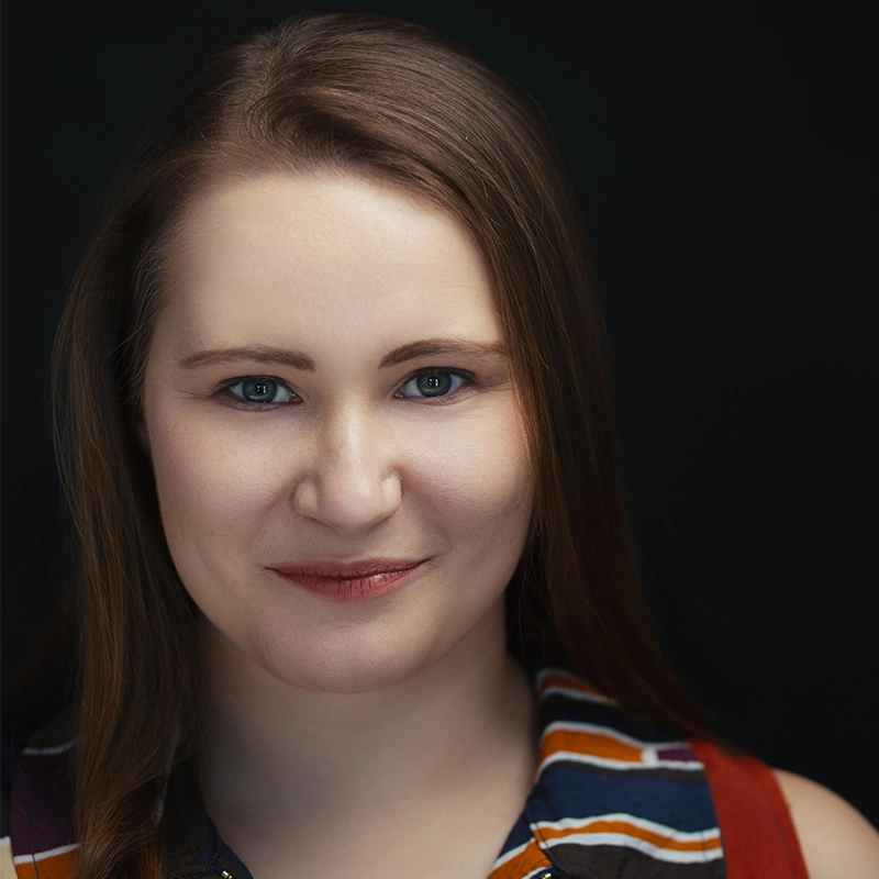 A woman with fair skin and straight, light brown hair smiles softly. Jenna Leigh Miller wears a sleeveless, striped shirt in orange, blue, and white. The plain black background puts focus on her warmly lit face.