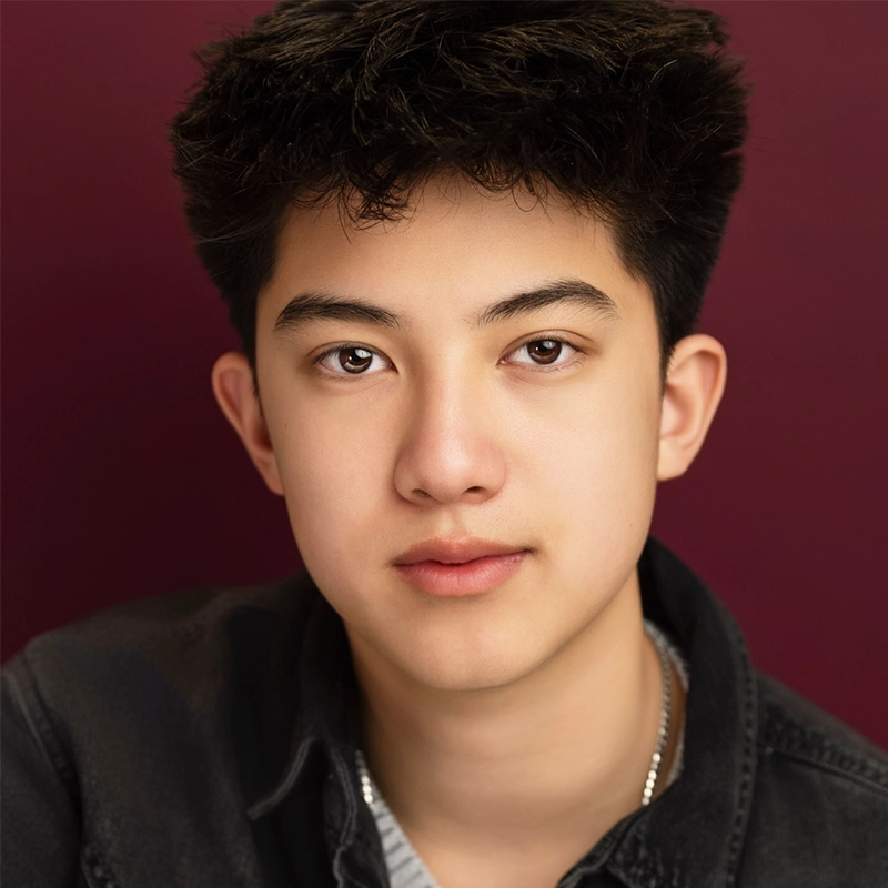 A young person with short, thick dark hair and fair skin looks directly at the camera. They have brown eyes, smooth skin, and wear a black collared shirt over a gray top. The background is a solid deep burgundy color.