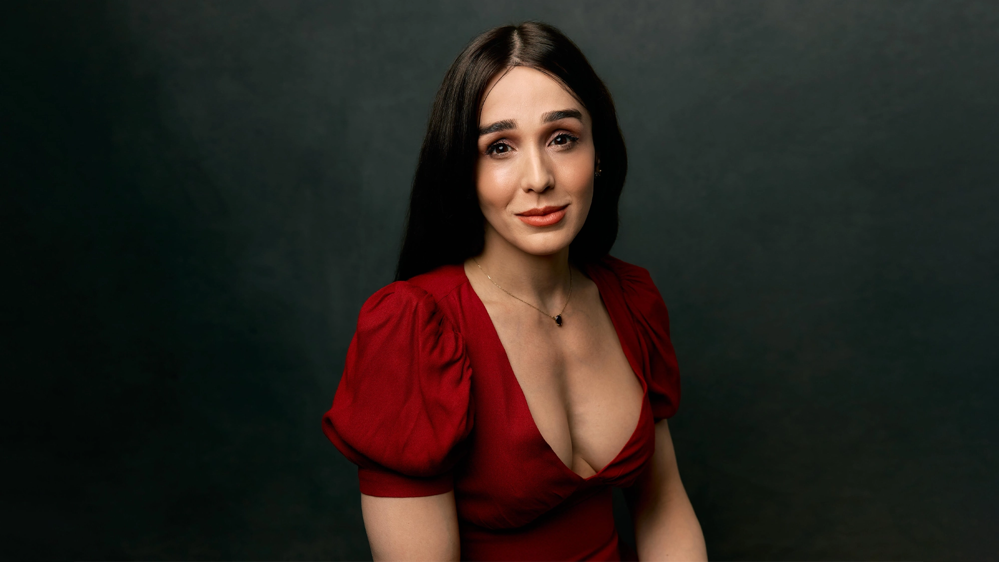 A woman with long dark hair and light skin sits against a dark background, wearing a red dress with puffed sleeves and a deep neckline. She has a gentle expression, natural makeup, and wears a small pendant necklace—evoking the soulful mood of 