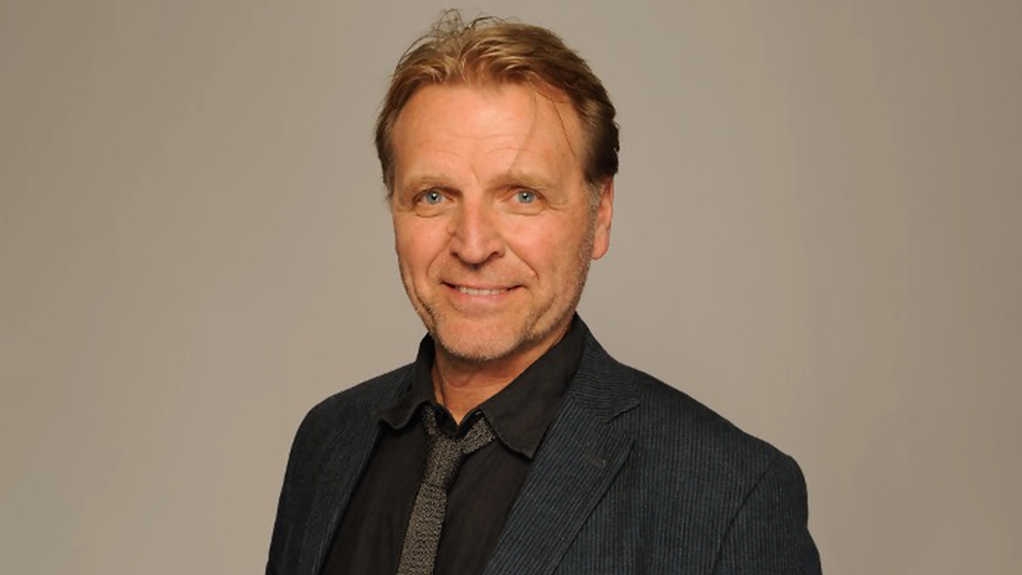 A middle-aged man with light hair and blue eyes smiles at the camera. He is dressed in a dark blazer, black shirt, and patterned tie. The even lighting highlights his friendly expression, reminiscent of David Rasche from the David Rasche show.
