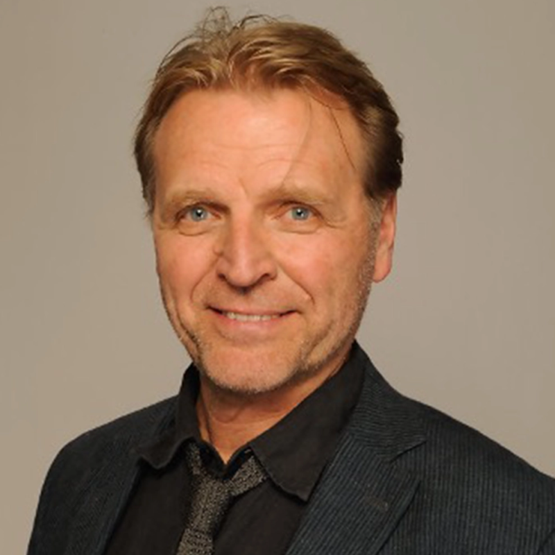A middle-aged man with light skin, blond hair, and blue eyes smiles at the camera. He is wearing a dark striped blazer over a black shirt and a patterned tie. The background is plain and light-colored, creating a professional portrait atmosphere.