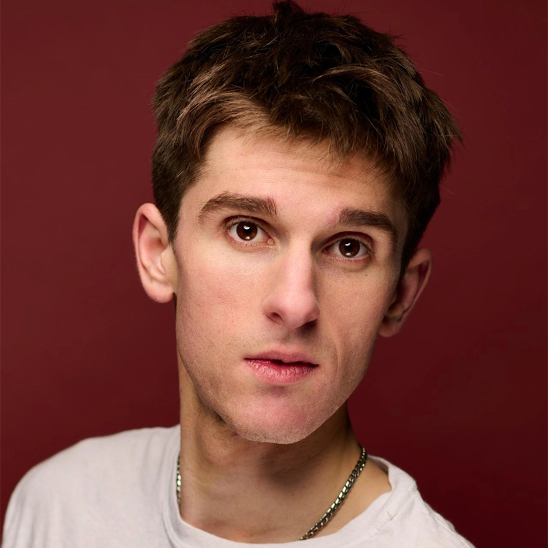 A young man with short brown hair and brown eyes looks slightly to the side against a deep red background. He wears a light gray t-shirt and a thin silver chain necklace. His expression is neutral, and the lighting highlights his facial features.