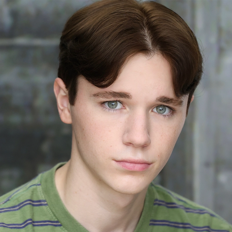 A young person with fair skin, light green eyes, and short brown hair parted in the middle looks directly at the camera with a neutral expression. Jordan Gulliford wears a green striped shirt and stands against a blurred, grayish background.