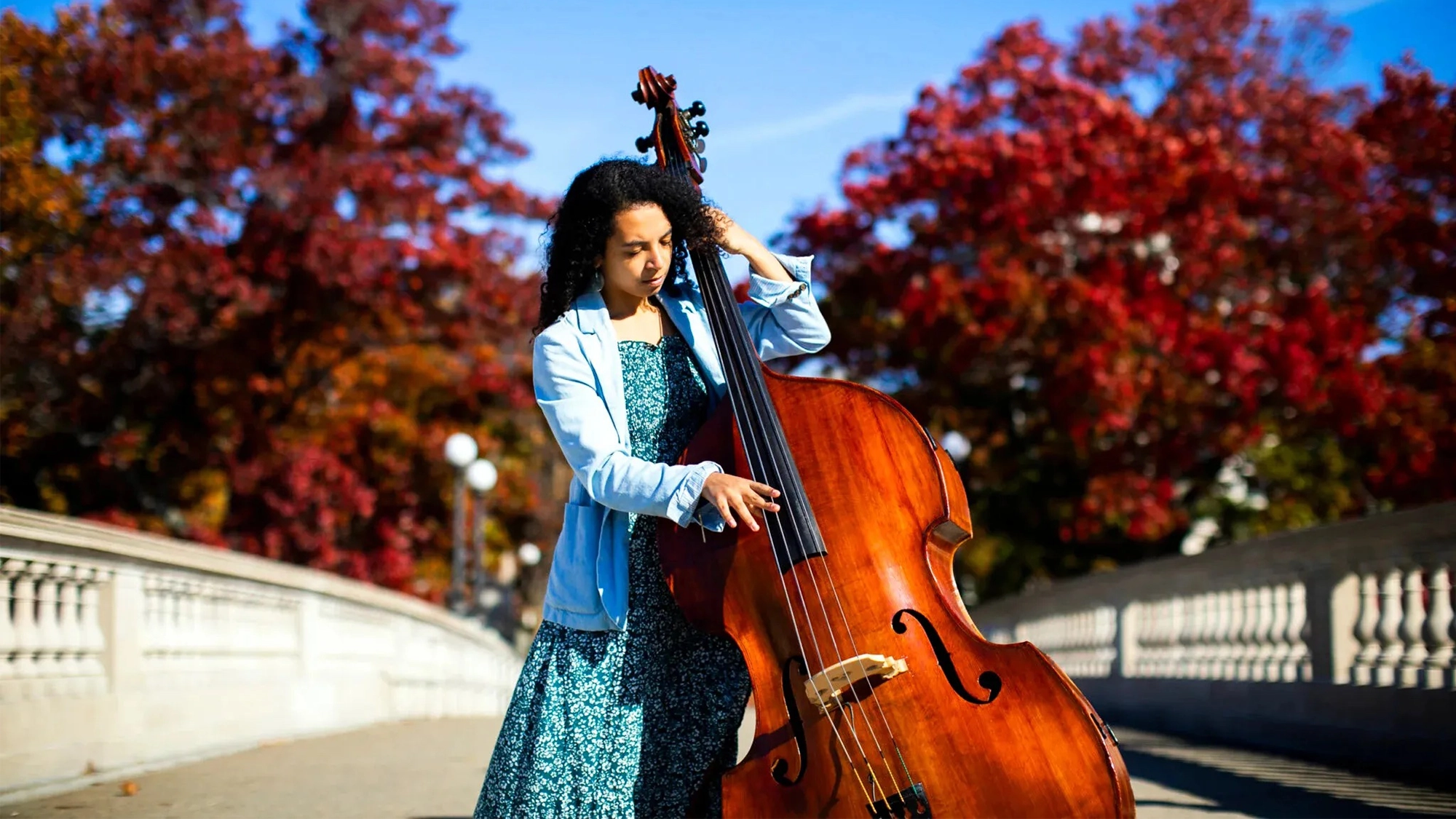 A woman in a blue dress and light jacket plays a double bass outdoors on a bright, sunny day. She stands on a wide walkway flanked by white railings, with vibrant red autumn trees and a clear blue sky framing her modern jazz performance.