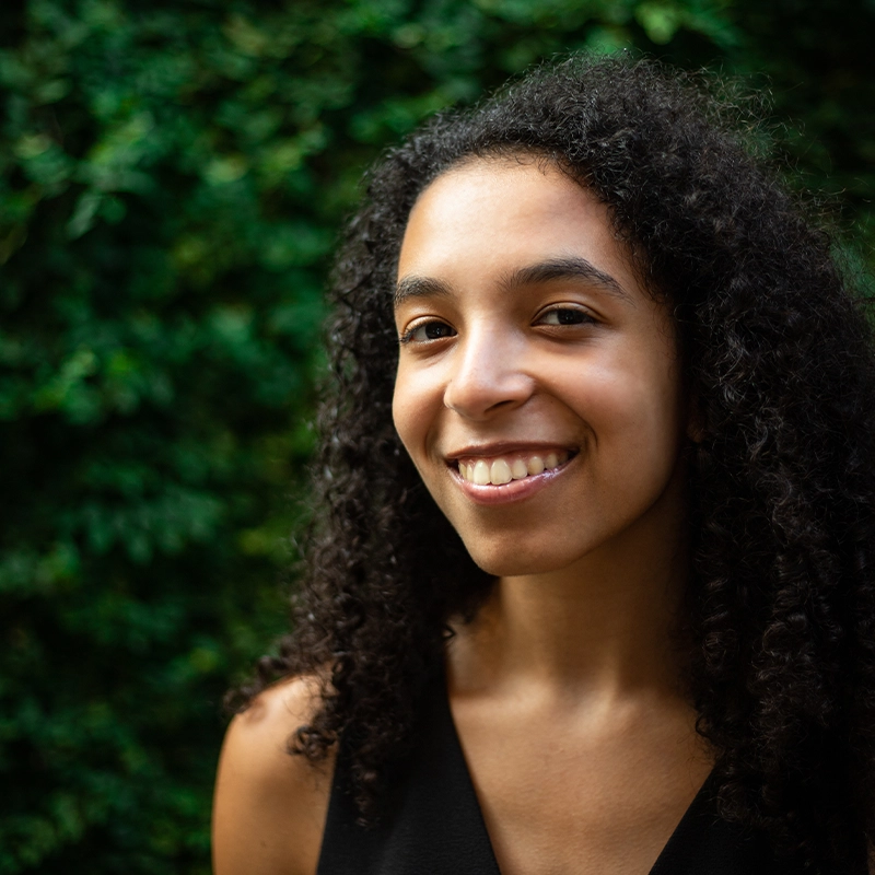 A young woman with curly dark hair smiles at the camera. She is wearing a sleeveless black top and stands outdoors in front of lush green foliage. The lighting is natural, and the mood is bright and friendly.