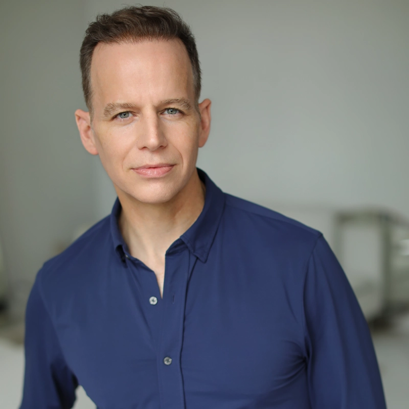James Donegan, a man with short brown hair and blue eyes, wearing a blue button-up shirt, stands in a softly lit, minimalistic indoor setting. He faces the camera with a neutral, slight smile and relaxed posture.
