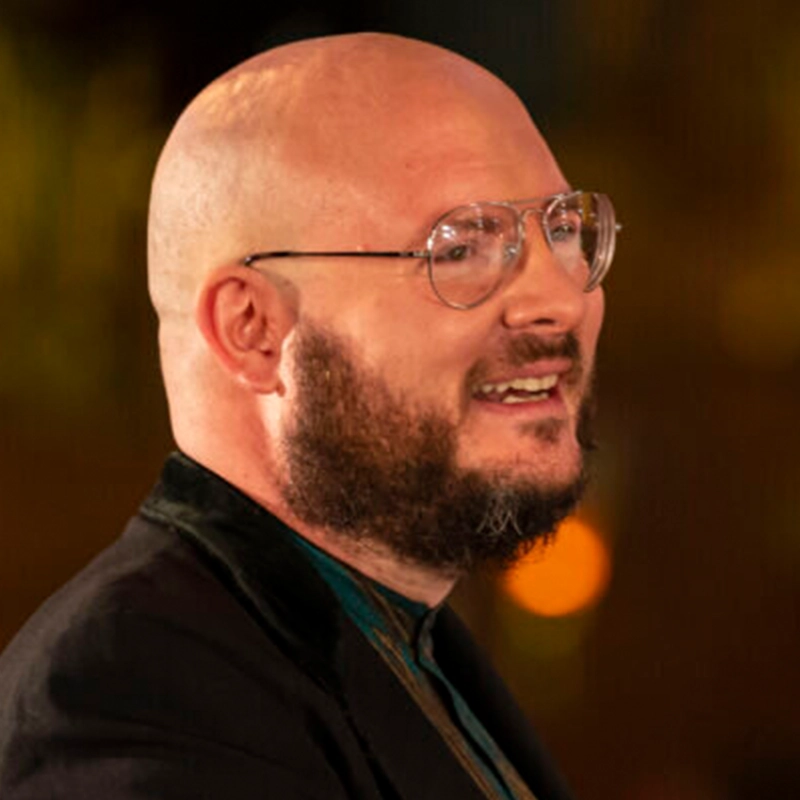 A bald man with a full beard and clear round glasses smiles while looking to the right. He wears a dark jacket over a green shirt. The background is softly blurred with warm yellow lights, suggesting an indoor or evening setting.