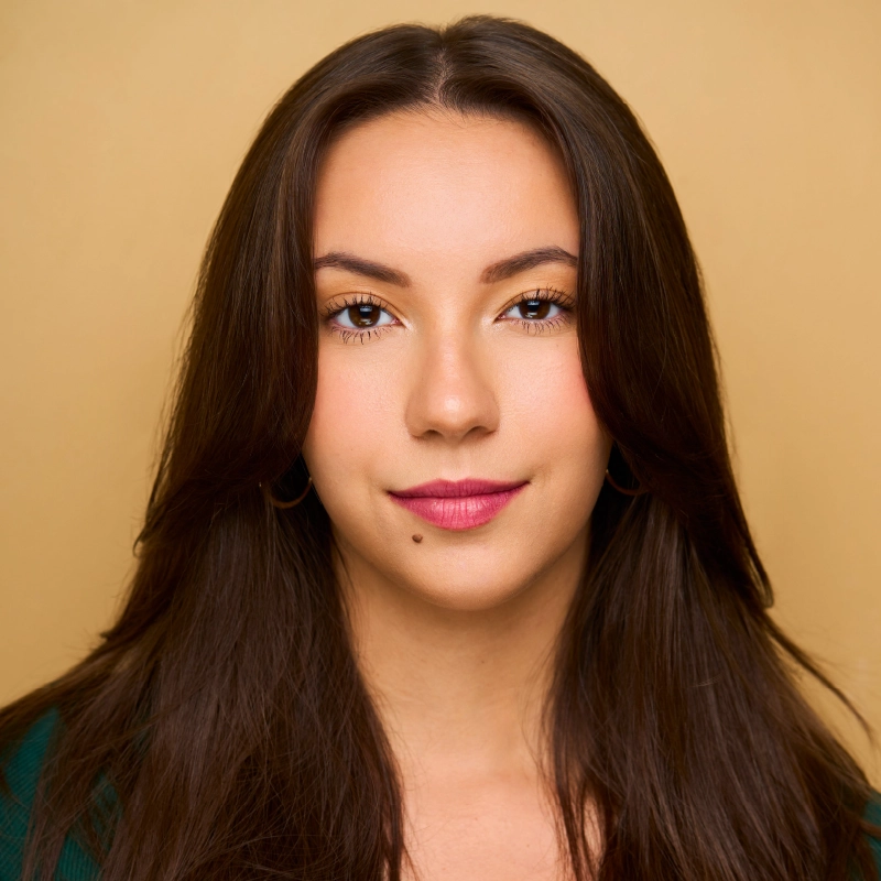 A young woman with long, straight brown hair and light skin, wearing a dark green top. She has hoop earrings, subtle makeup, bold pink lipstick, and a beauty mark above her lip. The background is a solid warm beige color. She gazes confidently at the camera.