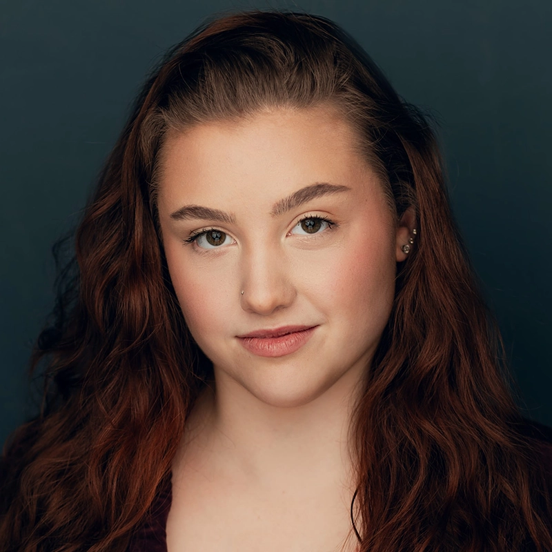 Jenna Leigh Miller, a young woman with long, wavy auburn hair and fair skin, looks confidently at the camera. She has hazel eyes, natural makeup, a small nose ring, and multiple ear piercings against a plain dark blue background.