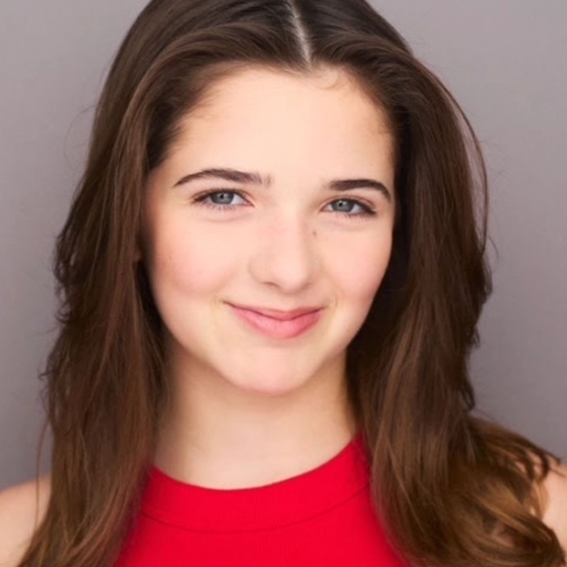 A young woman with long brown hair and blue eyes smiles gently at the camera. She is wearing a sleeveless, bright red top by Jake Hamilton and is positioned against a plain light gray background. Her expression is friendly and confident.