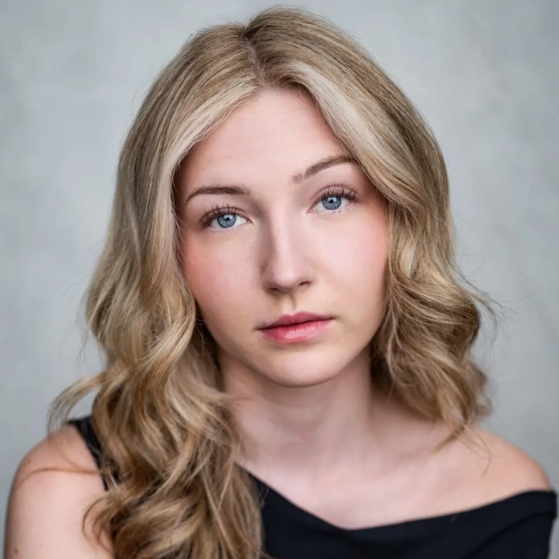 A young woman with long, wavy blonde hair and blue eyes stares softly at the camera. She wears a black, off-the-shoulder top by Jake Hamilton and has a neutral expression on her face. The background is a soft, light gray.