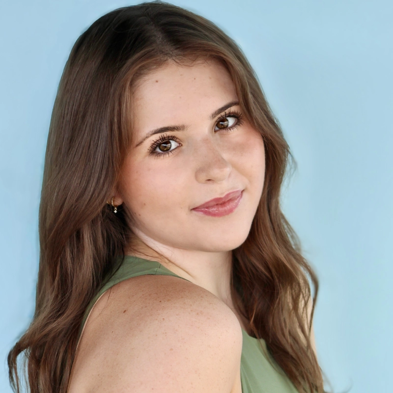 A young woman with long, wavy brown hair and fair skin smiles softly at the camera. She wears a sleeveless green top and stands against a light blue background. Her head is turned slightly over her shoulder, giving a gentle and relaxed expression.