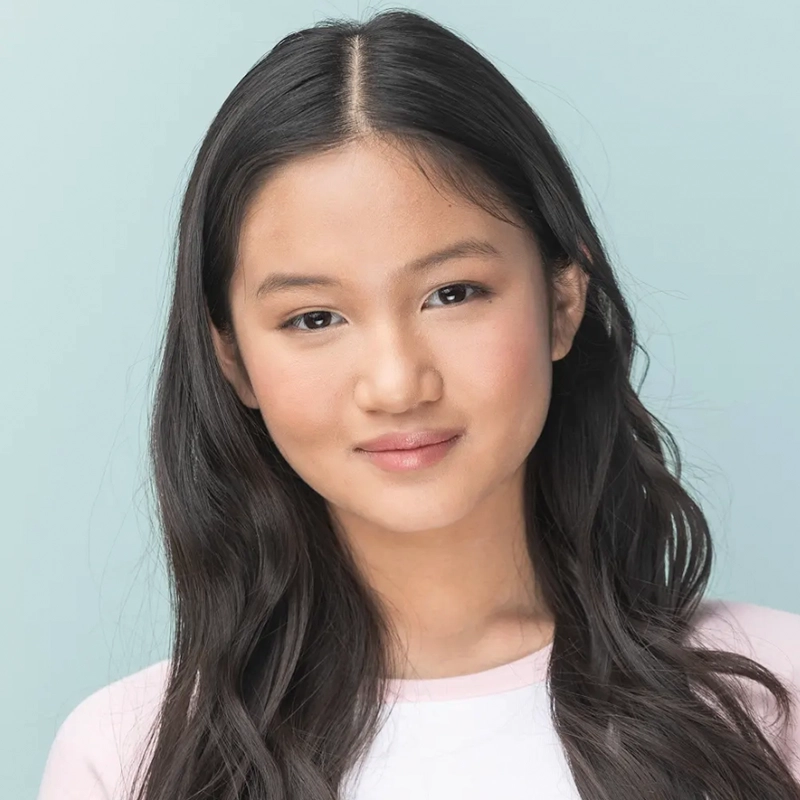 A young woman with long, wavy dark hair and smooth skin looks at the camera with a gentle smile. Styled by Jake Hamilton, she wears a white top with pale pink sleeves, standing confidently against a soft blue background.