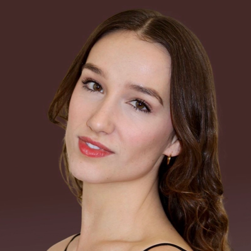 A young woman with long, wavy brown hair and fair skin poses in front of a plain brown background. She has light makeup, glossy lips, and is wearing a black spaghetti strap top and small gold hoop earrings, gazing softly at the camera.