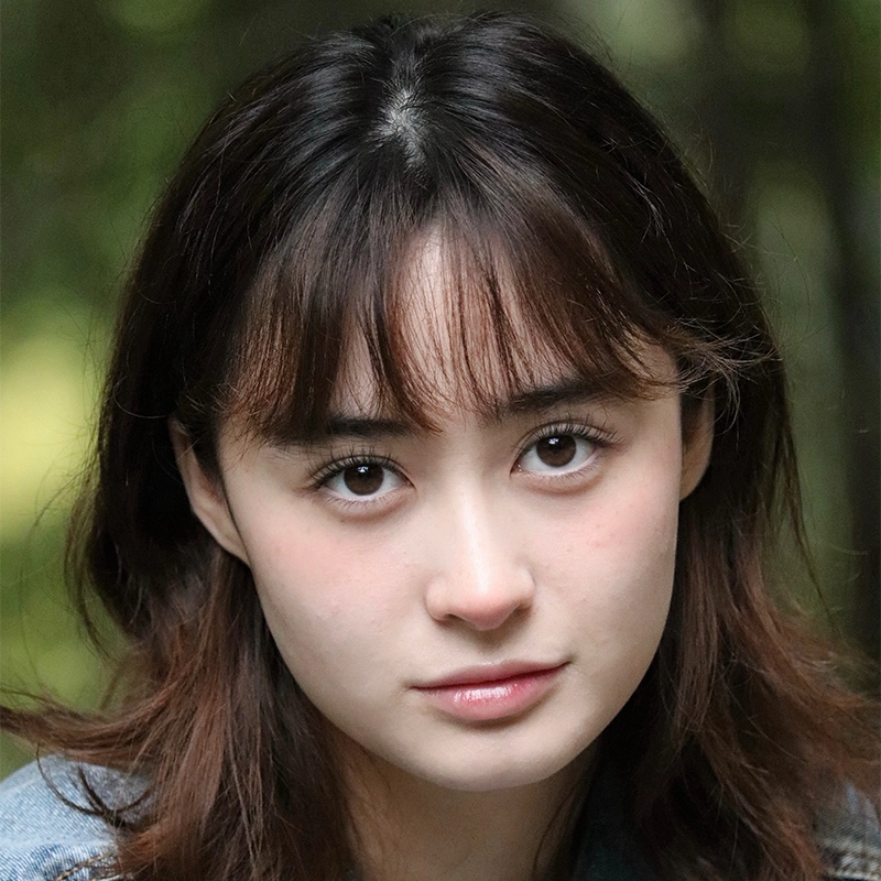 A young woman with fair skin and brown hair looks directly at the camera. She has fringe bangs, soft makeup, and brown eyes. The blurred greenery in the background suggests a serene outdoor setting, reminiscent of a Jake Hamilton film.