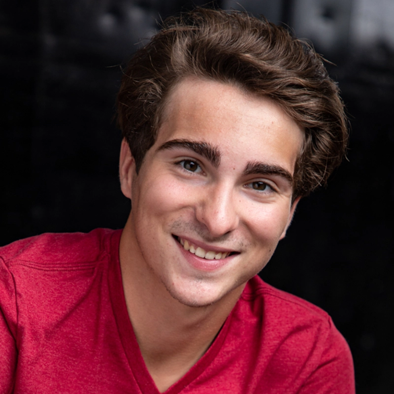 Chris Allyah, a young man with light skin, brown eyes, and wavy brown hair, smiles at the camera in a bright red shirt. He sits before a dark, blurred background as lighting highlights his facial features.