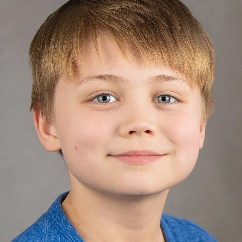 A young boy with straight light brown hair and blue eyes smiles softly. He is wearing a blue shirt and facing the camera against a plain gray background. His calm, friendly expression gives him a gentle, almost Charlemagne-like presence.