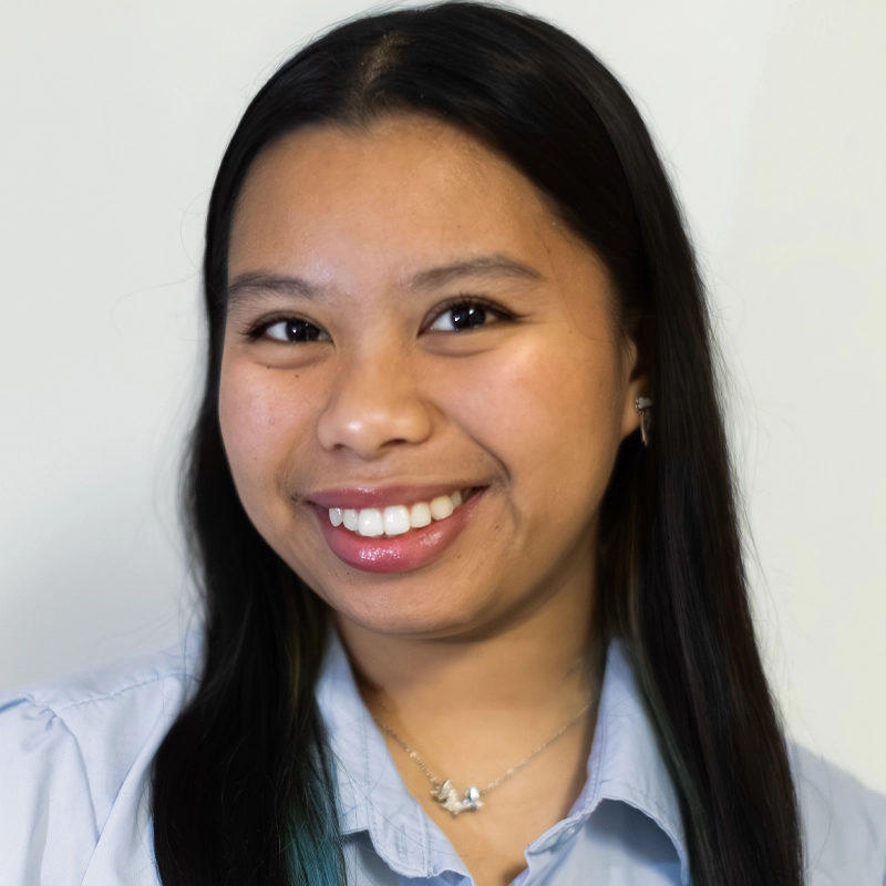 Noelle Flores, with long, straight black hair, smiles at the camera. She wears a light blue collared shirt and a silver necklace. The plain, light background keeps the focus on her warm and friendly expression.