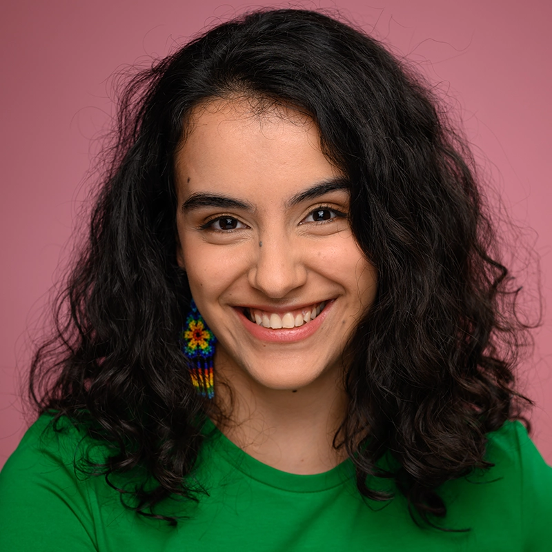 A young woman with medium skin tone, curly dark hair, and colorful beaded earrings smiles warmly. She wears a green shirt and is posed in front of a solid pink background.
