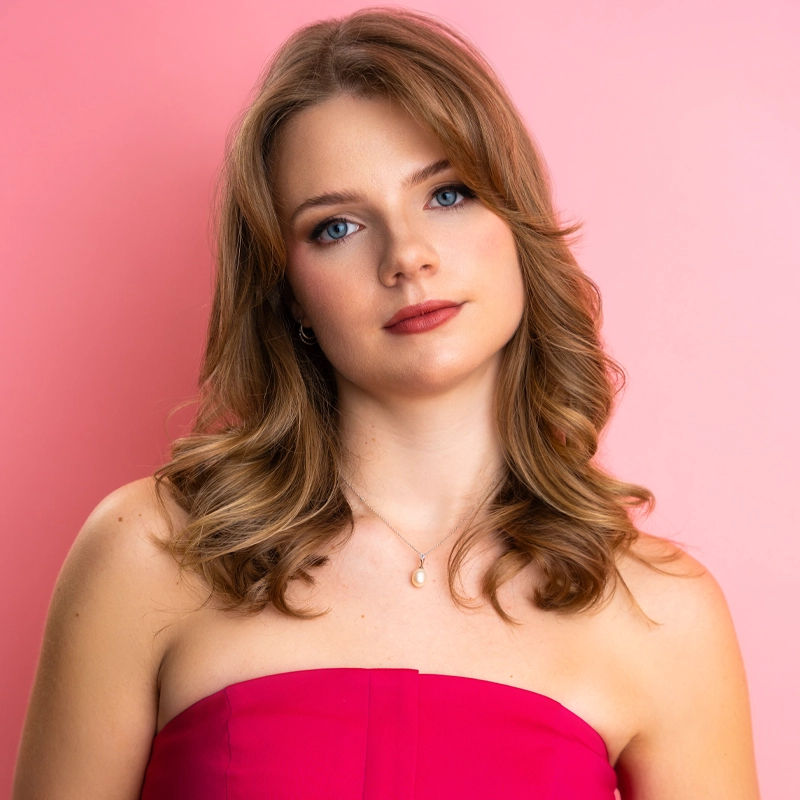 Allie George, a young woman with wavy light brown hair and blue eyes, poses in front of a pink background. She wears a strapless fuchsia top, a simple necklace with a small pendant, and natural makeup with soft pink lipstick.