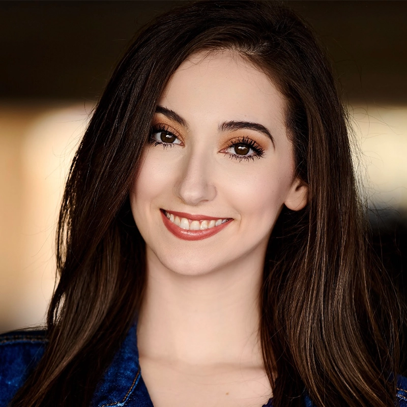 Gina Marie Incandela, a young woman with long brown hair and brown eyes, smiles at the camera. She has smooth, fair skin, natural makeup with defined brows and peach lipstick, wearing a blue denim jacket against a softly blurred warm background.