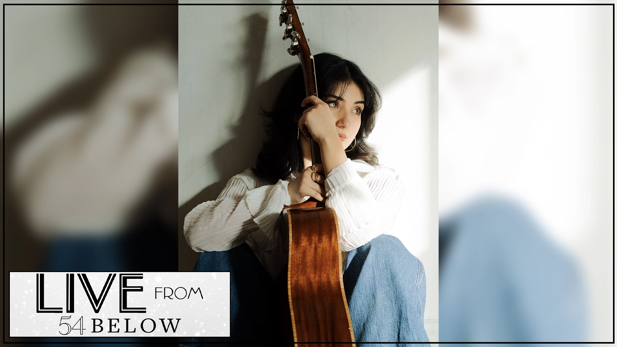 A young woman with dark hair sits against a wall holding an acoustic guitar. She wears a white shirt and blue jeans, looking thoughtfully to the side. The bottom left corner features 