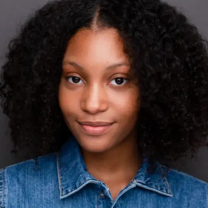 A young woman with medium brown skin and natural, shoulder-length curly hair smiles gently at the camera. She wears a blue denim shirt with the collar visible. The background is a plain, dark gray, drawing focus to her face in this Jake Hamilton portrait style.
