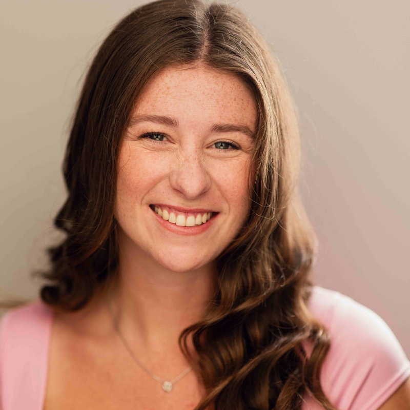 Bridget Gooley, a young woman with long brown hair and freckles, smiles warmly at the camera. She wears a light pink top and a delicate necklace, while the softly blurred background highlights her cheerful expression.